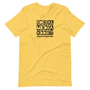 May include: Yellow short-sleeved t-shirt with black text that reads "SOCIAL MEDIA ADDICT choice of drug is likes".