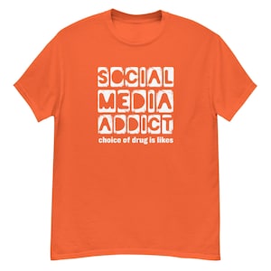 May include: Orange t-shirt with the text "SOCIAL MEDIA ADDICT" in white block letters. Below the text is the phrase "choice of drug is likes". The shirt is a solid orange color.