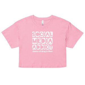 May include: A pink crop top with the words "SOCIAL MEDIA ADDICT" in a white, blocky font. Below, the text reads "choice of drug is likes". The shirt has a crew neck and short sleeves.