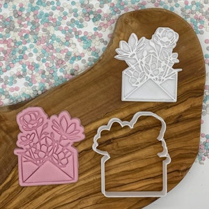 May include: Three cookie cutters shaped like flowers in an envelope. One is pink, one is white, and one is a white outline. The cutters are on a wooden surface with colourful sprinkles.