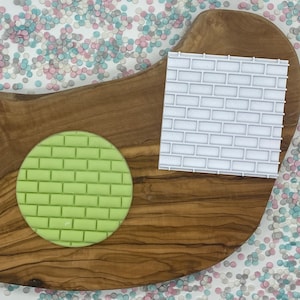 May include: A light green circular brick pattern and a white square brick pattern are displayed on a wooden cutting board. The brick patterns are used for decorating cakes and cookies. The background has a scattering of pastel confetti.