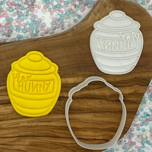 May include: Three honey pot-shaped cookie cutters. One is yellow, with the word "HUNNY" on it. Another is white, and the third is an outline of the pot. The cutters are on a wooden board, with a scattering of colorful confetti.