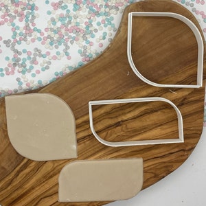 May include: A wooden chopping board with three white plastic cookie cutters in various shapes. Two pieces of tan dough are cut to match the cutters. Multi-coloured sprinkles are scattered in the background.