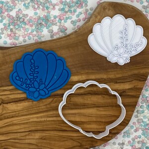 May include: Three seashell-shaped cookie cutters in blue and white, with floral details. One blue cutter is on the left, a white cutter is on the top right, and a white frame-shaped cutter is at the bottom. The cutters are on a wooden surface.