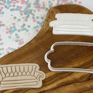 May include: Three white plastic cookie cutters shaped like a sofa, set against a wooden board with colourful sprinkles. The sofa designs vary, with one having a detailed back.