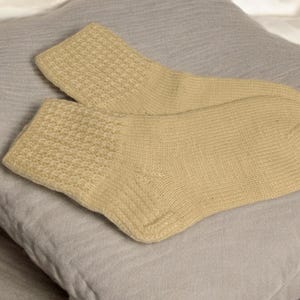 May include: A pair of light yellow socks with textured cuffs, resting on a gray pillow. The socks are made of a soft, knitted material, and the cuffs have a detailed pattern. The background is a soft, neutral color.