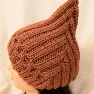 Puede incluir: Un gorro tejido a mano de color óxido con una forma única y alargada. El gorro presenta un diseño de punto de cable texturizado y una parte superior puntiaguda. El gorro está hecho de un hilo suave y cálido, perfecto para el clima frío. El gorro se muestra en una cabeza de maniquí.