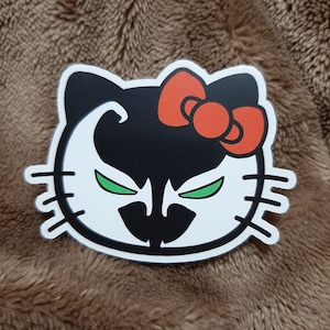 May include: A white Hello Kitty sticker with a black and green Spawn character design. The character has a red bow on top of its head.