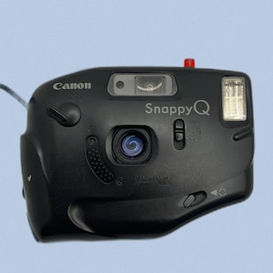 May include: Black Canon Snappy Q 35mm film camera. The camera has a built-in flash, a lens, and a viewfinder. The camera has the Canon logo on the top left and the Snappy Q logo on the top centre.