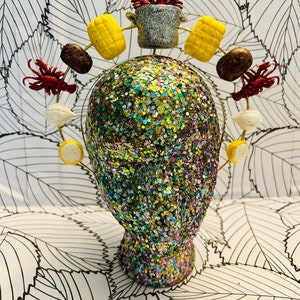 May include: A silver pot with red crawfish on top, surrounded by yellow corn, brown potatoes, and white onions. The pot is on a headband with a glittery background.