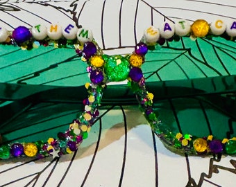 May include: Green, purple, and gold glitter sunglasses with beads spelling "Let Them Eat Cake" on the frame.