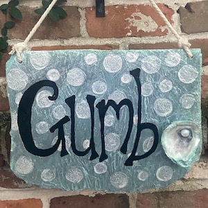 May include: A blue and white painted wooden sign with the word "Gumbo" written in black. The sign has a white pearl in a shell on the right side.