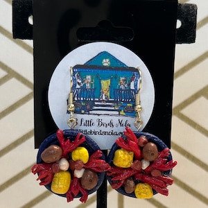 May include: A pair of blue and red clay earrings shaped like a plate of crawfish, corn, and potatoes. The earrings are handmade and feature a gold hook.