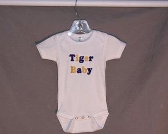May include: White baby onesie with the text "Tiger Baby" in navy blue and gold lettering. The onesie has short sleeves and snaps at the bottom. It is hanging on a clear plastic hanger.