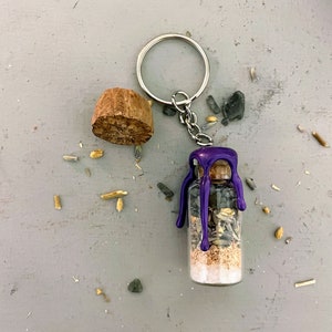 Puede incluir: Una pequeña botella de vidrio con un sello de cera púrpura, llena de hierbas secas y cristales, cuelga de un llavero plateado. La botella está unida a un tapón de corcho.