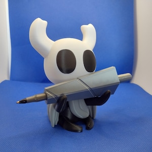 May include: A white and black figurine of a character holding a grey pen. The figurine has large black eyes and two curved horns. The pen is grey and black. The figurine is on a blue surface.