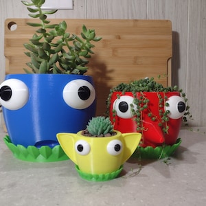 May include: Three 3D printed planters shaped like faces with googly eyes. The planters are blue, red, and yellow and each has a different succulent plant growing in it.