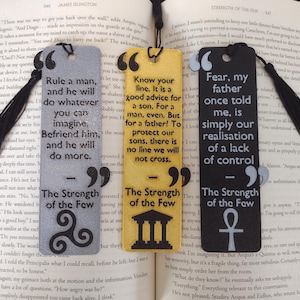 Peut inclure: Trois marque-pages rectangulaires avec des citations de "The Strength of the Few" sont affich&eacute;s sur un livre ouvert. Les marque-pages sont gris, jaunes et noirs, chacun avec un pompon noir. Le texte est en blanc.