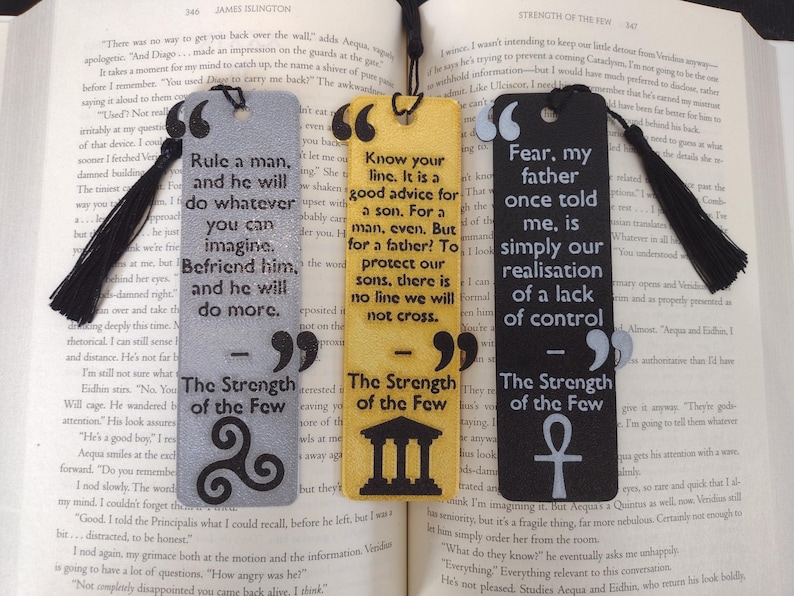 Peut inclure: Trois marque-pages rectangulaires avec des citations de "The Strength of the Few" sont affich&eacute;s sur un livre ouvert. Les marque-pages sont gris, jaunes et noirs, chacun avec un pompon noir. Le texte est en blanc.