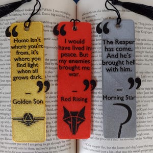 May include: Three rectangular bookmarks with quotes from books. One is gold with black text, one is red with a black wolf symbol, and one is gray with a black scythe. Each has a black tassel.