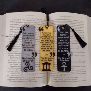 Peut inclure: Trois marque-pages rectangulaires avec des citations et des pompons noirs. Les marque-pages sont gris, jaunes et noirs, chacun avec une citation diff&eacute;rente de "The Strength of the Few". Le marque-page gris a un n&oelig;ud celtique, le jaune un b&acirc;timent et le noir un symbole ankh.