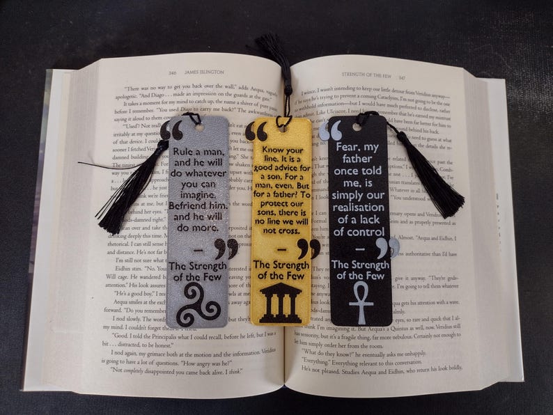 Peut inclure: Trois marque-pages rectangulaires avec des citations et des pompons noirs. Les marque-pages sont gris, jaunes et noirs, chacun avec une citation diff&eacute;rente de "The Strength of the Few". Le marque-page gris a un n&oelig;ud celtique, le jaune un b&acirc;timent et le noir un symbole ankh.