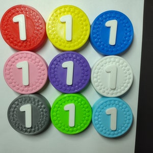 Peut inclure: Neuf jetons ronds en plastique de couleurs différentes avec le chiffre "1" imprimé en blanc sur chacun. Les jetons sont disposés en trois rangées de trois.
