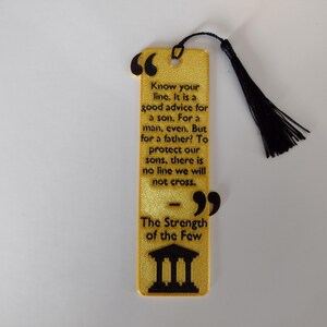 Peut inclure: Un marque-page dor&eacute; avec du texte noir et un pompon noir. Le marque-page contient une citation sur la famille et la force, avec les mots "The Strength of the Few" en bas. Le marque-page est rectangulaire avec des bords arrondis.