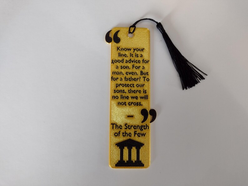 Peut inclure: Un marque-page dor&eacute; avec du texte noir et un pompon noir. Le marque-page contient une citation sur la famille et la force, avec les mots "The Strength of the Few" en bas. Le marque-page est rectangulaire avec des bords arrondis.