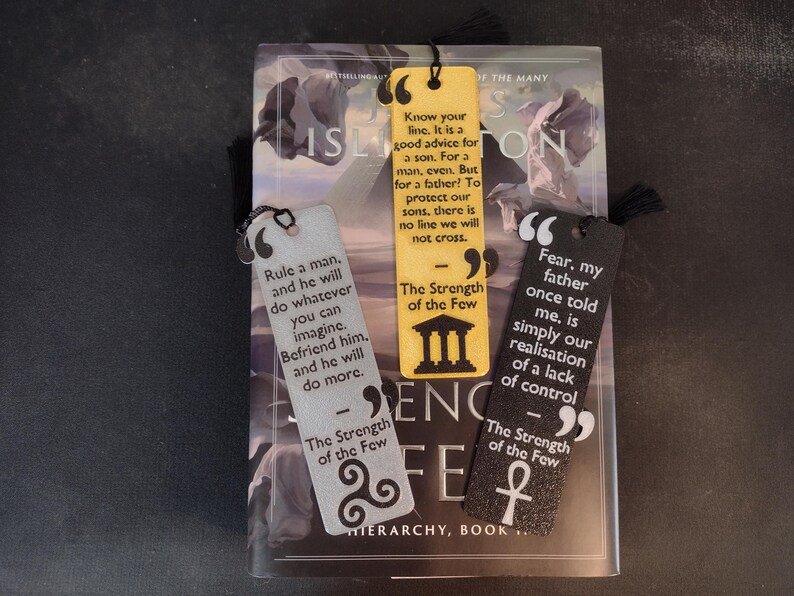 Peut inclure: Trois marque-pages d&eacute;coratifs avec des citations de "The Strength of the Few" sont pr&eacute;sent&eacute;s sur un livre. Les marque-pages sont jaunes, argent&eacute;s et noirs, chacun avec un design unique et un pompon. Le titre du livre est "ISLINGTON".
