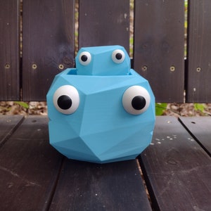 Könnte beinhalten: Ein hellblauer geometrischer Blumentopf mit großen, weißen Glitz Augen. Der Topf hat einen kleineren, hellblauen geometrischen Topf im Inneren, ebenfalls mit Glitz Augen.