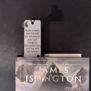 Peut inclure: Un marque-page argent&eacute; avec un pompon noir, portant la citation &laquo;&nbsp;Gouvernez un homme, et il fera tout ce que vous pouvez imaginer. Soyez son ami, et il en fera plus.&nbsp;&raquo; Le marque-page est pos&eacute; sur un livre de James Islington.