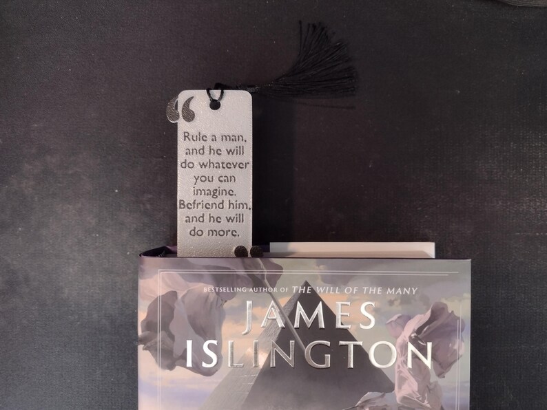 Peut inclure: Un marque-page argent&eacute; avec un pompon noir, portant la citation &laquo;&nbsp;Gouvernez un homme, et il fera tout ce que vous pouvez imaginer. Soyez son ami, et il en fera plus.&nbsp;&raquo; Le marque-page est pos&eacute; sur un livre de James Islington.