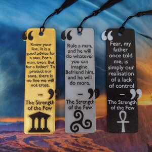 Peut inclure: Trois marque-pages rectangulaires avec des pompons noirs. Les marque-pages sont dor&eacute;s, argent&eacute;s et noirs, chacun avec une citation et un symbole. Le marque-page dor&eacute; contient une citation sur la famille, l'argent&eacute; sur la manipulation et le noir sur la peur.