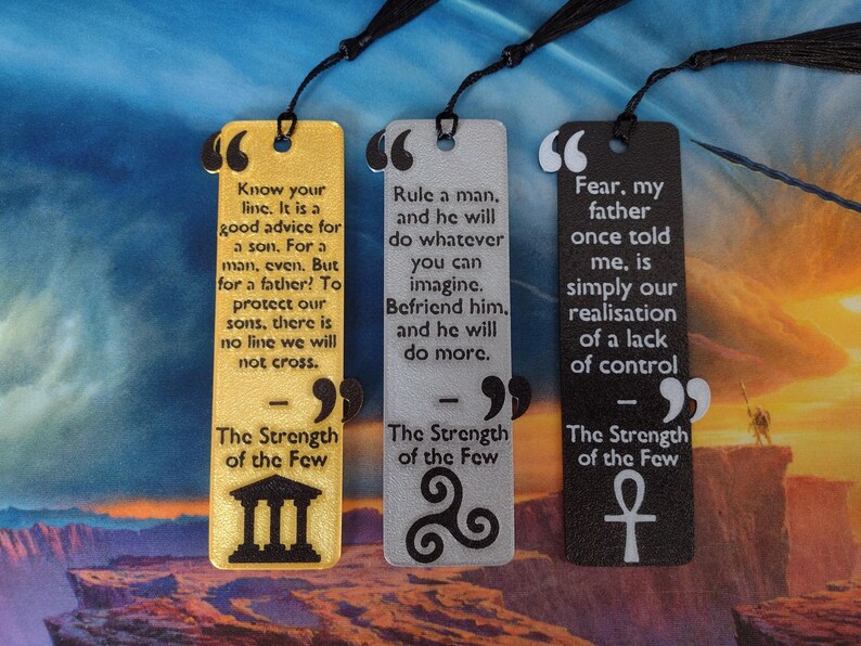 Peut inclure: Trois marque-pages rectangulaires avec des pompons noirs. Les marque-pages sont dor&eacute;s, argent&eacute;s et noirs, chacun avec une citation et un symbole. Le marque-page dor&eacute; contient une citation sur la famille, l'argent&eacute; sur la manipulation et le noir sur la peur.