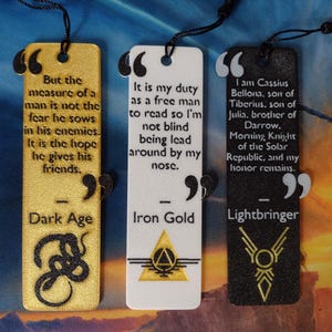 May include: Three rectangular bookmarks with quotes and designs. One is gold with a snake, the second is white with a triangle, and the third is black with a geometric design. Each has a black cord.