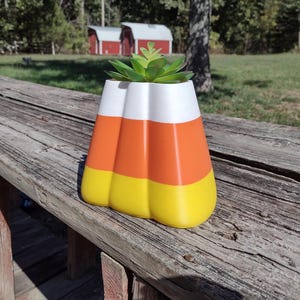 Puede incluir: Una maceta decorativa con forma de caramelo de maíz, con secciones blancas, naranjas y amarillas. La maceta contiene una pequeña planta suculenta verde. La maceta está sobre una superficie de madera envejecida.