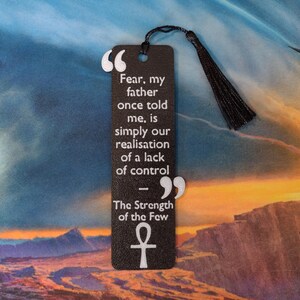 Peut inclure: Un marque-page noir avec une citation sur la peur et le contr&ocirc;le, avec du texte blanc et un pompon. Le marque-page comprend le symbole ankh et la citation "La peur, mon p&egrave;re m'a dit un jour, est simplement notre prise de conscience d'un manque de contr&ocirc;le."