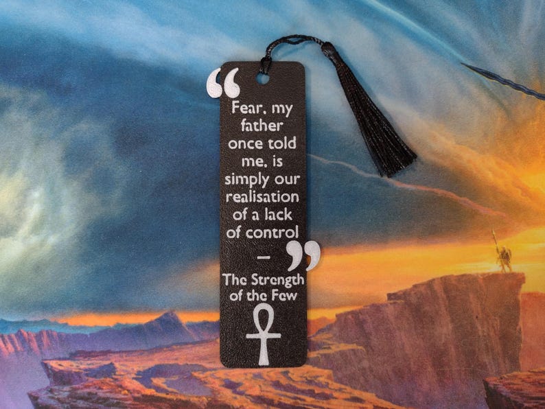 Peut inclure: Un marque-page noir avec une citation sur la peur et le contr&ocirc;le, avec du texte blanc et un pompon. Le marque-page comprend le symbole ankh et la citation "La peur, mon p&egrave;re m'a dit un jour, est simplement notre prise de conscience d'un manque de contr&ocirc;le."