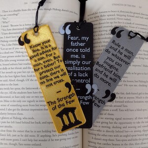 Peut inclure: Trois marque-pages d&eacute;coratifs avec des citations. L'un est dor&eacute; avec du texte noir, un autre est noir avec du texte blanc et le troisi&egrave;me est gris avec du texte noir. Chaque marque-page a un pompon noir.