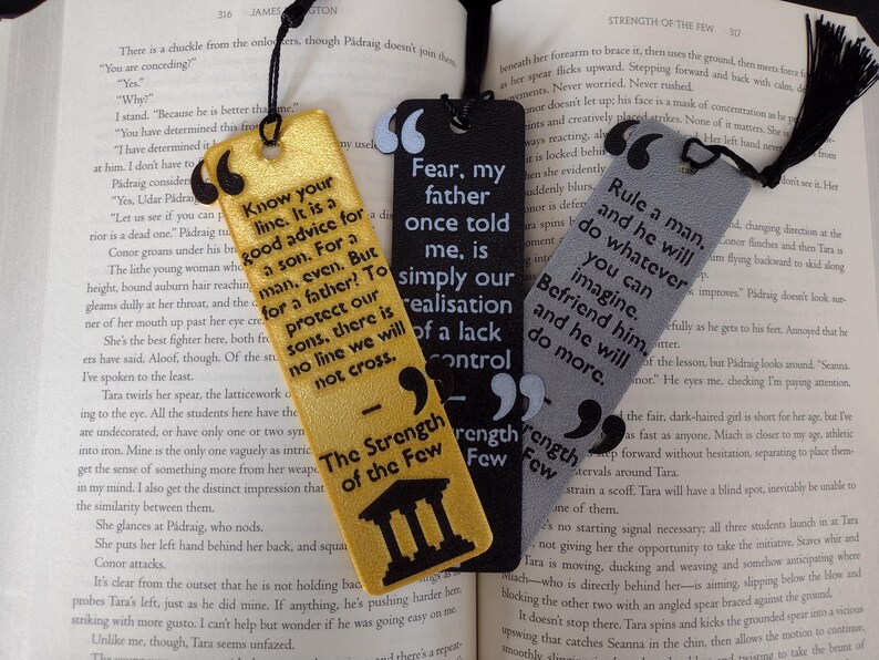Peut inclure: Trois marque-pages d&eacute;coratifs avec des citations. L'un est dor&eacute; avec du texte noir, un autre est noir avec du texte blanc et le troisi&egrave;me est gris avec du texte noir. Chaque marque-page a un pompon noir.