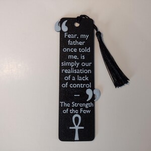 Peut inclure: Marque-page noir avec un pompon, avec une citation en texte blanc : "La peur, mon p&egrave;re m'a dit un jour, est simplement notre prise de conscience d'un manque de contr&ocirc;le." Comprend un symbole ankh et le texte "La force des quelques-uns."
