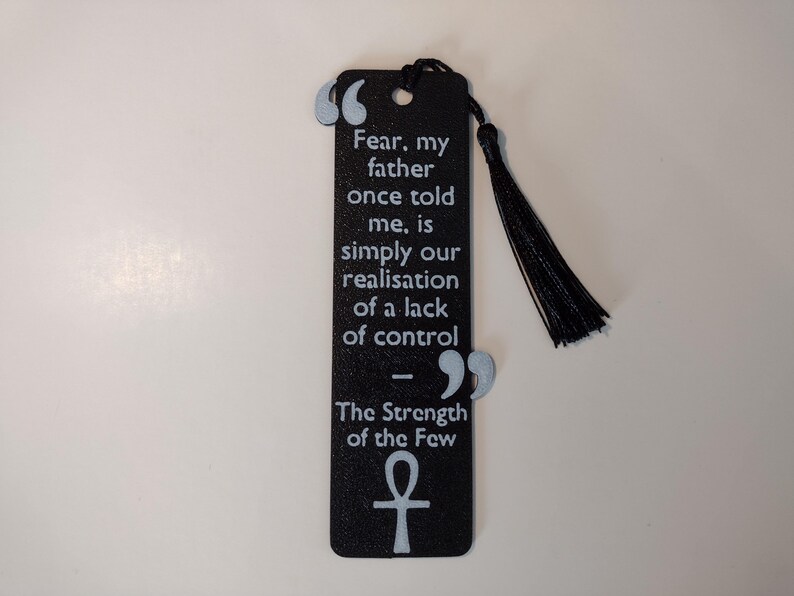 Peut inclure: Marque-page noir avec un pompon, avec une citation en texte blanc : "La peur, mon p&egrave;re m'a dit un jour, est simplement notre prise de conscience d'un manque de contr&ocirc;le." Comprend un symbole ankh et le texte "La force des quelques-uns."