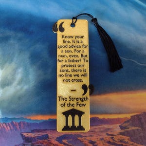 Peut inclure: Marque-page rectangulaire en bois avec un pompon noir. Le marque-page porte le texte grav&eacute; : "Know your line...The Strength of the Few" et un dessin de b&acirc;timent. Le fond est un paysage color&eacute;.
