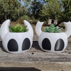 May include: Two white, stylized planters with black eye-shaped details and curved handles, each holding a green succulent plant. The planters are set on a wooden surface, with a blurred background of trees and foliage.