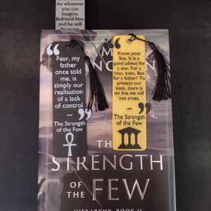 Peut inclure: Trois marque-pages avec des pompons noirs reposent sur un livre intitul&eacute; "The Strength of the Few". Les marque-pages pr&eacute;sentent des citations et des motifs en noir, blanc et jaune. La couverture du livre est sombre avec des lettres argent&eacute;es.