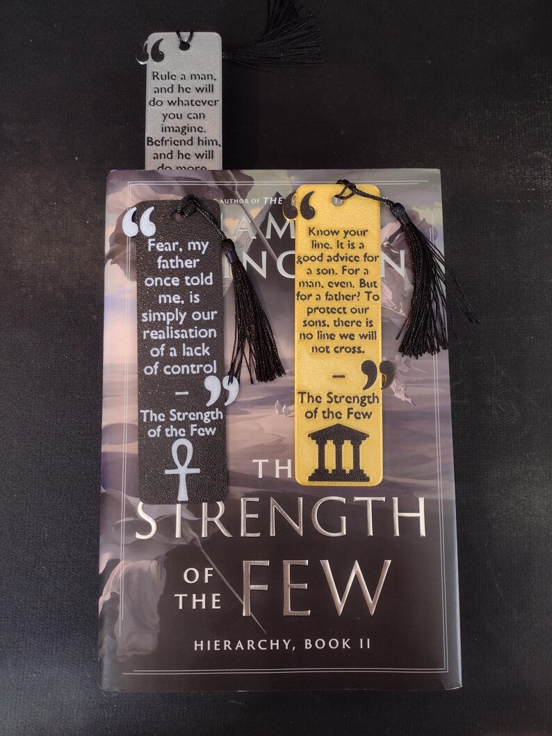 Peut inclure: Trois marque-pages avec des pompons noirs reposent sur un livre intitul&eacute; "The Strength of the Few". Les marque-pages pr&eacute;sentent des citations et des motifs en noir, blanc et jaune. La couverture du livre est sombre avec des lettres argent&eacute;es.
