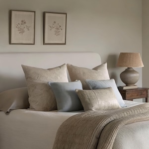 Puede incluir: Un dormitorio con una cama adornada con cojines en tonos beige y azul claro, una manta tejida y una colcha a juego. Dos grabados bot&aacute;nicos enmarcados cuelgan sobre la cama. Una l&aacute;mpara con pantalla tejida descansa sobre una mesita de noche de madera.