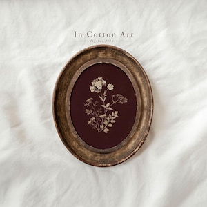 Puede incluir: Una impresión enmarcada ovalada con una ilustración botánica de flores y follaje blancos sobre un fondo burdeos oscuro. El marco tiene un acabado dorado envejecido. El texto "In Cotton Art digital prints" está en la parte superior.
