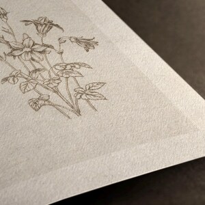 Puede incluir: Ilustraci&oacute;n detallada de un colibr&iacute; y elementos florales, incluyendo flores y hojas, impresa en tinta marr&oacute;n sobre un papel texturizado blanquecino. La esquina del papel es visible sobre un fondo oscuro.